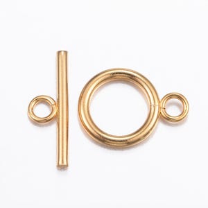 Può includere: Chiusura a toggle dorata, composta da un anello circolare con un piccolo anello e una barra dritta con un anello. La chiusura è progettata per la creazione di gioielli, su sfondo bianco.