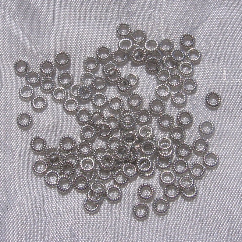 May include: A large pile of silver jump rings, a type of jewelry finding, on a white fabric background.