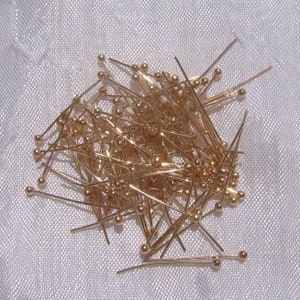 May include: A pile of gold-colored sewing pins with small ball heads. The pins are scattered across a white, textured surface, creating a dense cluster of metallic elements.