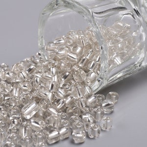 May include: A glass jar filled with silver seed beads, spilling out onto a gray surface.