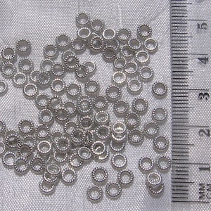 May include: A close-up of a pile of silver jump rings, measuring approximately 3 mm in diameter, against a white fabric background. A ruler is visible in the background.