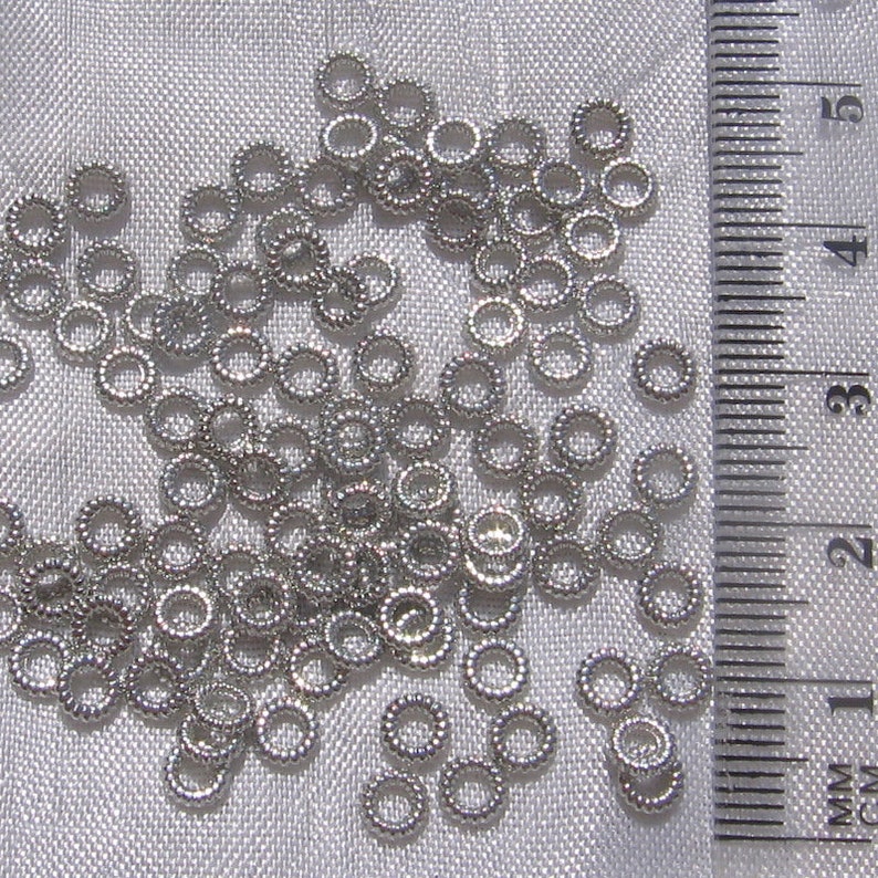 May include: A close-up of a pile of silver jump rings, measuring approximately 3 mm in diameter, against a white fabric background. A ruler is visible in the background.
