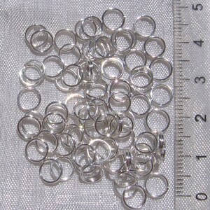 May include: A pile of silver jump rings, approximately 1/2 inch in diameter, against a white fabric background. A ruler is visible in the background, with markings from 0 to 5.
