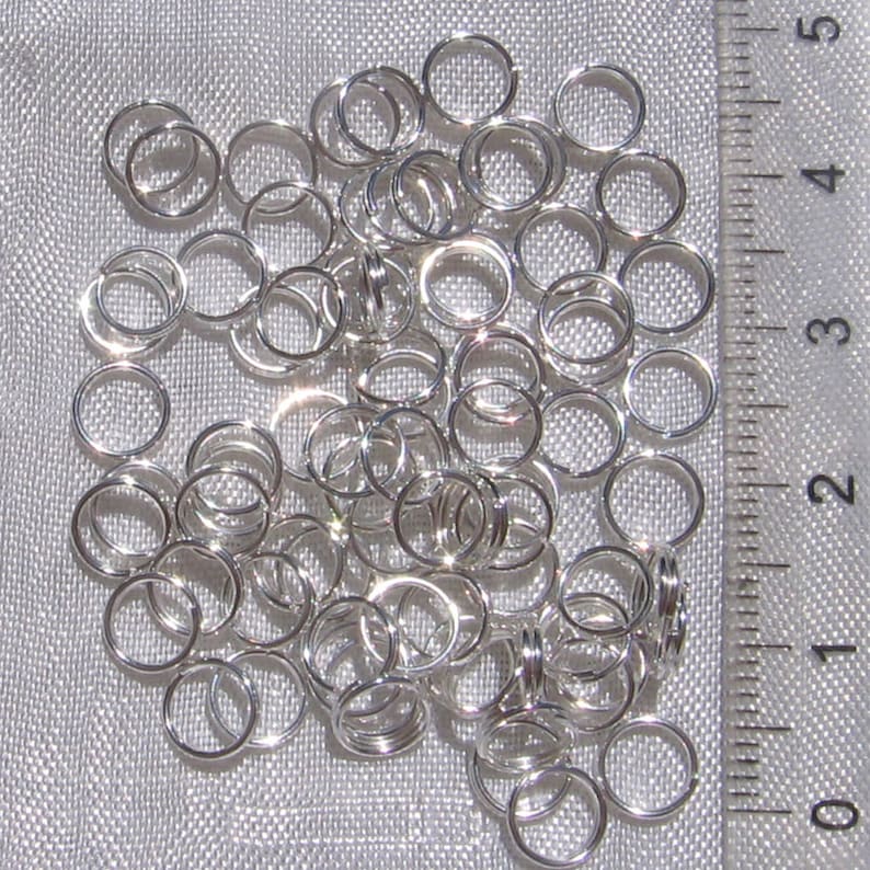 May include: A pile of silver jump rings, approximately 1/2 inch in diameter, against a white fabric background. A ruler is visible in the background, with markings from 0 to 5.