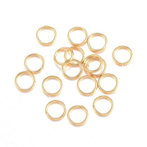 May include: A group of 15 gold jump rings, scattered on a white surface. The jump rings are small and circular, with a smooth, shiny finish.