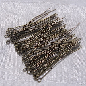 Può includere: Un mucchio di forcine per capelli color bronzo con estremità ad anello, disposte su una superficie bianca e strutturata. Le forcine sono sottili e dritte, con una leggera curva vicino all'estremità ad anello. Accessori per capelli metallici.