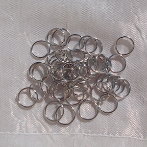 Peut inclure: Une collection de nombreux petits anneaux de jonction métalliques circulaires de couleur argentée. Chaque anneau individuel présente une ouverture fendue unique, permettant une fixation et une connexion faciles pour la bijouterie ou divers projets d'artisanat.
