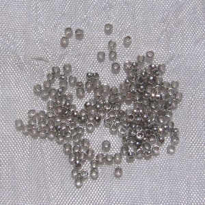 May include: A large quantity of small, silver-colored metal beads. The beads are round and have a hole in the center. They are scattered on a white fabric surface.