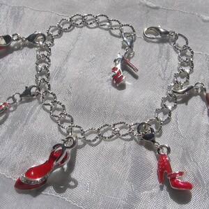 K&ouml;nnte beinhalten: Ein silberfarbenes Bettelarmband mit mehreren h&auml;ngenden Anh&auml;ngern in Form von hochhackigen Schuhen. Jeder Schuh-Anh&auml;nger ist mit einer leuchtend roten Emaille-Beschichtung und silbernen Akzenten versehen. Die Kette besteht aus texturierten Gliedern.