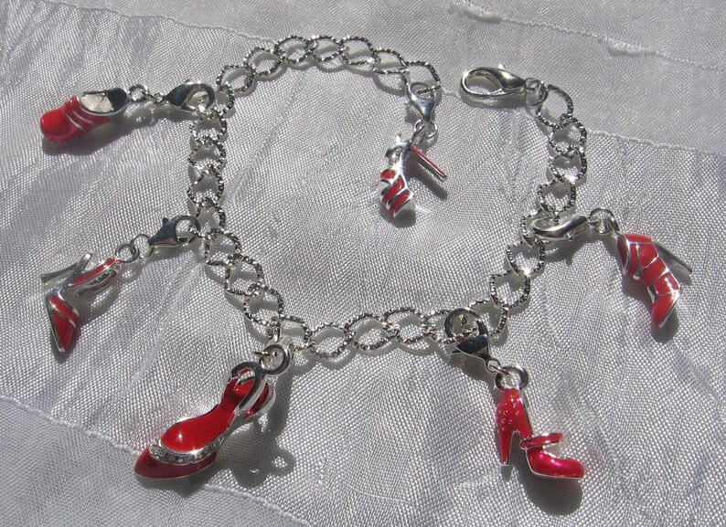 K&ouml;nnte beinhalten: Ein silberfarbenes Bettelarmband mit mehreren h&auml;ngenden Anh&auml;ngern in Form von hochhackigen Schuhen. Jeder Schuh-Anh&auml;nger ist mit einer leuchtend roten Emaille-Beschichtung und silbernen Akzenten versehen. Die Kette besteht aus texturierten Gliedern.