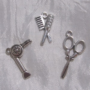 May include: Three silver-toned metal charms shaped like hairdressing tools: a hair dryer, a comb and brush, and a pair of scissors.