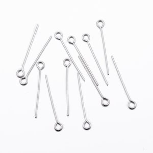 May include: A group of silver head pins with eye loops. The pins are scattered on a white surface.