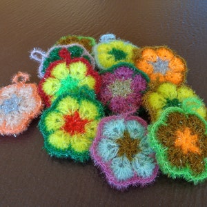 Könnte beinhalten: Eine Sammlung bunter, gehäkelter Blumen-Spüllappen in den Farben Grün, Gelb, Orange, Rosa und Blau. Die Spüllappen sind aus einem flauschigen Garn gefertigt und haben eine glitzernde Oberfläche.