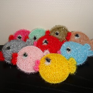 Könnte beinhalten: Eine Gruppe bunter gehäkelter Fisch-Ornamente mit glitzerndem Garn. Die Fische sind in verschiedenen Farben, darunter rosa, blau, grün, rot, silber und gelb.