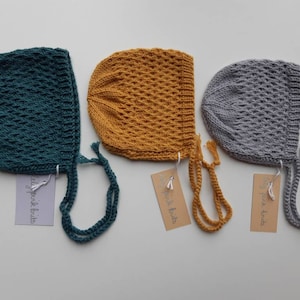 May include: Three crocheted baby bonnets in teal, mustard yellow, and gray with matching ties. The bonnets have a diamond stitch pattern and are labeled "Lily Pad Knits".