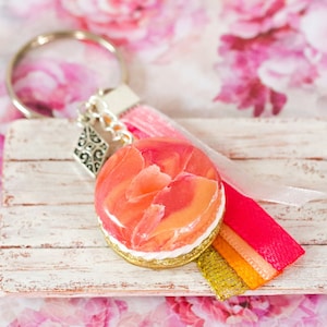 May include: A pink and orange floral keychain with a white wooden background. The keychain has a silver charm and a pink, orange, and gold ribbon.