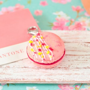 Könnte beinhalten: Ein rosa Macaron mit weißem Zuckerguss und gelben und rosa Streuseln auf einem weißen Holztablett mit dem Wort "PANTONE" auf einer rosa Karte.
