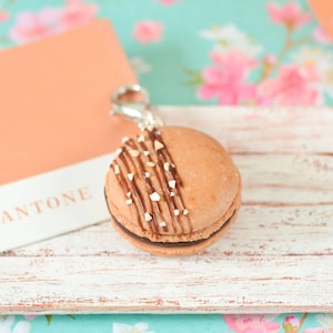 May include: A brown and white macaron with chocolate drizzle and white sprinkles on top. The macaron is attached to a silver key chain and is sitting on a white wooden tray. The tray is on a floral background.