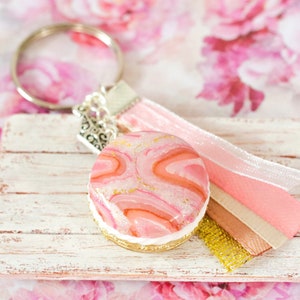 Könnte beinhalten: Rosa-goldfarbener Marmor-Macaron-Schlüsselanhänger mit silbernem Anhänger und rosa-goldenen Schleifenakzenten.