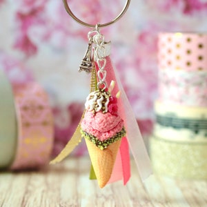 May include: A pink and white ice cream cone keychain with a green sprinkle topping, a white chocolate swirl, and a red raspberry. The keychain also features a silver Eiffel Tower charm and a pink, white, and green ribbon.