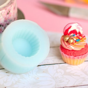 May include: A light blue silicone cupcake mould with a single cavity. A pink and white cupcake with gold sprinkles and a red and white candy on top is next to the mould.