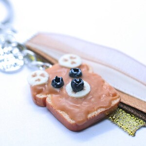 May include: A brown and tan bread keychain with a peanut butter and banana filling and two blueberries on top. The keychain is attached to a silver key ring with a charm that says "Live, Love, Laugh".