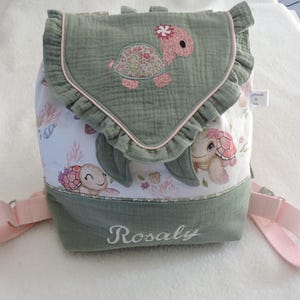 Può includere: Uno zaino verde e bianco con un design a tartaruga. La patta presenta un'applicazione a tartaruga con un fiore rosa e un bordo a balze. Lo zaino ha spallacci rosa e il nome "Rosaly" ricamato sul davanti.