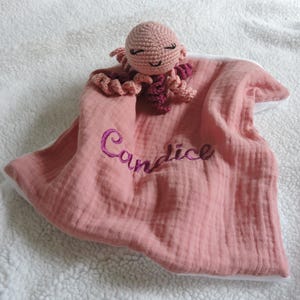 Peut inclure: Une couverture de sécurité rose pour bébé avec un jouet poulpe crocheté. Le nom "Candice" est brodé en violet sur la couverture. La pieuvre est rose clair avec des tentacules rose foncé.