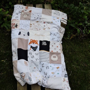 Peut inclure: Une courtepointe en patchwork réalisée à partir de vêtements de bébé avec différents motifs d'animaux, dont des pandas, des renards et des moutons. La courtepointe est beige, blanche et noire avec les mots "Le Petit Champion" et "Hello" brodés dessus.