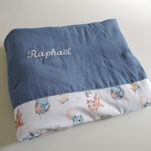 Peut inclure: Une couverture pour bébé bleue avec le nom "Raphaël" brodé en blanc. Le bord inférieur présente un tissu blanc avec un motif de dragons de dessins animés, de ballons et de lunes.