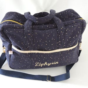 Può includere: Una borsa da viaggio blu navy con dettagli in glitter dorati e un nome ricamato in oro "Zéphyrin". La borsa ha una lunga tracolla blu navy e una cerniera color crema.