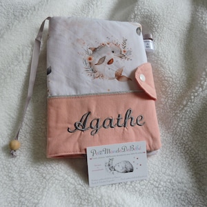 May include: A pink and white fabric book cover with a dolphin design and the name "Agathe" embroidered on the front. The cover has a flap with a button closure and a small tag that says "Petit Monde De Bebe".