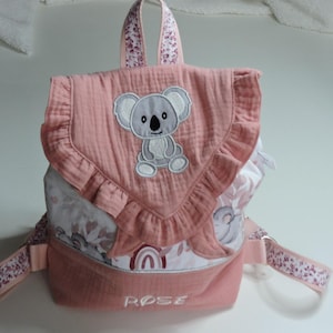 May include: A pink backpack with a koala bear applique. The backpack has a ruffled flap and floral patterned straps and handle. The word "ROSE" is embroidered on the front. The backpack is made of a soft, textured fabric.