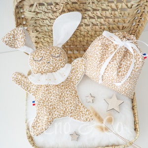Pode incluir: Um brinquedo de pelúcia de coelho bege e branco com um colar de renda branco e um padrão de folhas. O brinquedo está em uma cesta de vime com um forro de tecido branco. Há duas pequenas estrelas de madeira e uma pequena bolsa branca com um cordão branco.