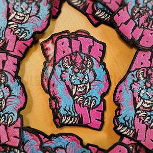 Może przedstawiać: Naszywka haftowana w kolorze różowym i niebieskim z tygrysem i napisem "BITE" czarnymi literami.