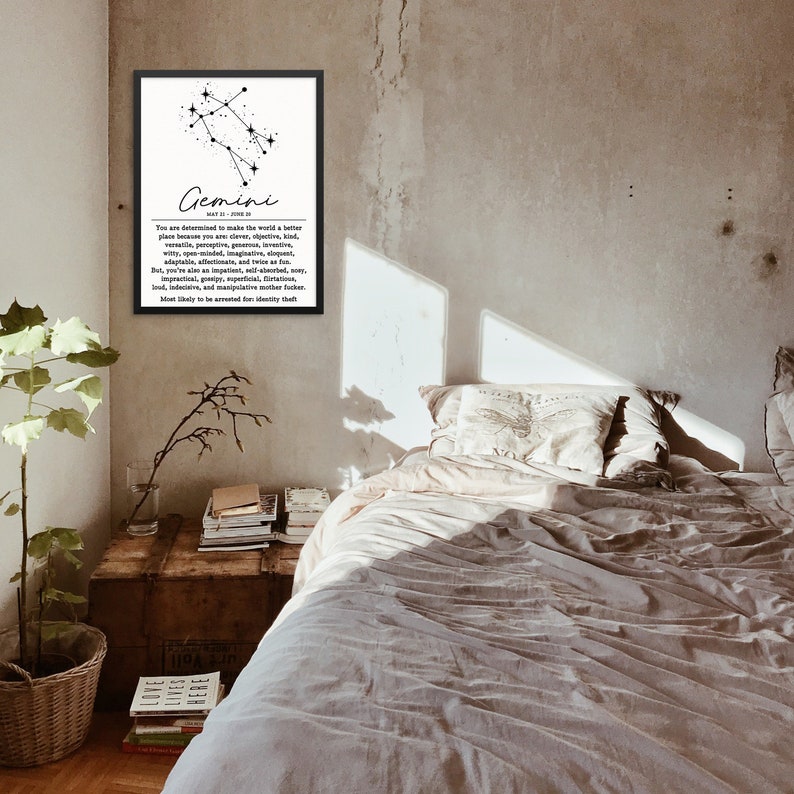 May include: A black and white framed print with the zodiac sign Gemini and a description of the Gemini personality traits. The print is hanging on a wall in a bedroom with a bed, pillows, and a side table with books.