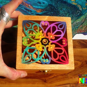 Op de afbeelding: Een houten doos met een regenboogkleurig, ingewikkeld uitgesneden ontwerp op de deksel. Het ontwerp heeft een bloemmotief met gouden accenten. De doos heeft een klein slotje aan de voorkant.
