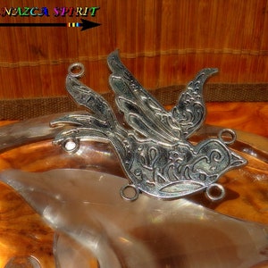 Può includere: Un ciondolo a forma di uccello in metallo argentato con dettagli intricati, tra cui ali aperte e motivi decorativi. Il ciondolo ha diversi anelli per il fissaggio a una collana o altri gioielli. L'immagine include anche il testo "Atelier NAZCA SPIRIT".