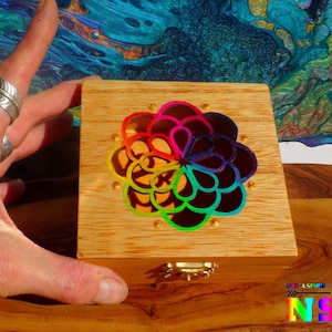 Op de afbeelding: Een kleine houten kist met een scharnierend deksel. De kist heeft een kleurrijk, regenboogkleurig bloemmotief op de bovenkant. De kist is gemaakt van lichtgekleurd hout en heeft een kleine, goudkleurige sluiting. Het Nazca Spirit-logo is rechtsonder zichtbaar.