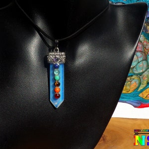 Peut inclure: Un collier pendentif avec un cristal bleu clair et sept perles de pierres précieuses colorées. Le pendentif est suspendu à un cordon noir et exposé sur un mannequin noir. Le collier a un dessus en métal argenté.