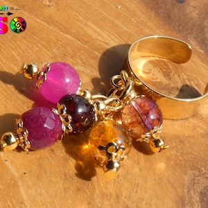 Op de afbeelding: Een goudkleurige verstelbare ring met een cluster van edelsteenbedels. De bedels omvatten gefacetteerde roze, bordeauxrode en amberkleurige kralen, geaccentueerd met goudkleurige metalen doppen en connectoren. De ring wordt gepresenteerd op een houten oppervlak.