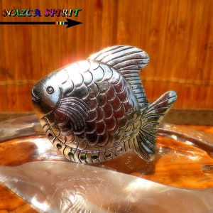 Può includere: Un oggetto decorativo a forma di pesce color argento con dettagli di squame e pinne. L'oggetto è appoggiato su una superficie riflettente. Il testo "Atelier Nazca Spirit" è visibile in alto a sinistra.
