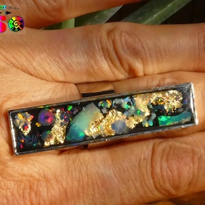 Peut inclure: Une bague rectangulaire avec un anneau argenté. La face de la bague présente un fond noir avec des pierres irisées incrustées, des flocons d'or et une touche de vert. La bague est tenue par une main.