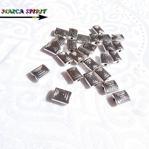 Peut inclure: Des perles rectangulaires argentées avec un motif géométrique sont dispersées sur une surface blanche. Les perles ont une forme carrée avec un motif en creux. L'image comprend également le texte "Atelier NAZCA SPIRIT" avec une flèche.
