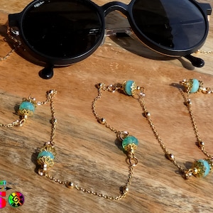 Può includere: Occhiali da sole rotondi neri con una catena dorata per occhiali. La catena presenta piccole perle dorate e perle decorative color turchese. Gli occhiali da sole e la catena sono esposti su una superficie di legno.