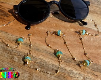 Könnte beinhalten: Schwarze runde Sonnenbrille mit einer goldenen Brillenbandkette. Die Kette hat kleine goldene Perlen und türkisfarbene Zierperlen. Die Sonnenbrille und die Kette werden auf einer Holzoberfläche präsentiert.