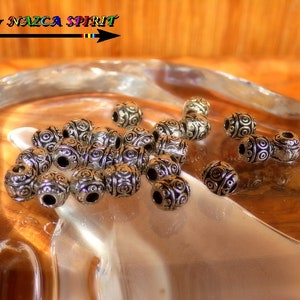 Op de afbeelding: Een close-up van een stapel zilveren metalen kralen met een draaiend patroon. De kralen liggen op een lichtbruin houten oppervlak.