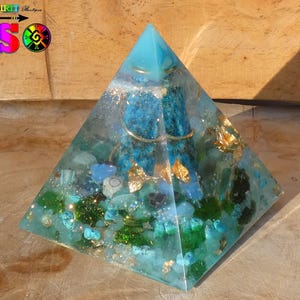 Pode incluir: Uma pirâmide de cristal azul cheia de vários cristais, incluindo turquesa, aventurina verde e folha de ouro. A pirâmide está sobre uma superfície de madeira.