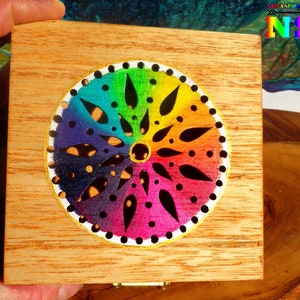 Puede incluir: Una caja de madera con un diseño de mandala arcoíris cortado en la tapa. El mandala está pintado con un degradado de colores, incluyendo morado, azul, verde, amarillo, naranja y rojo. La caja es una gran manera de guardar objetos pequeños, como joyas o baratijas.