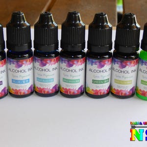 Può includere: Sette piccoli flaconi di inchiostro all'alcool in vari colori, tra cui blu zaffiro, blu, blu acquamarina, verde mela, verde, verde limone e verde limone. Ogni flacone ha un'etichetta che recita "Alcohol Ink" con il nome del colore stampato sotto.